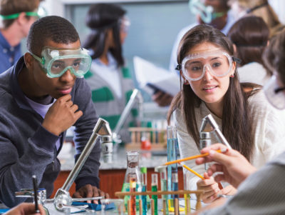 Career Academies: two high school students wearing goggles and listening to a teacher who points a pencil at some chemistry equipment