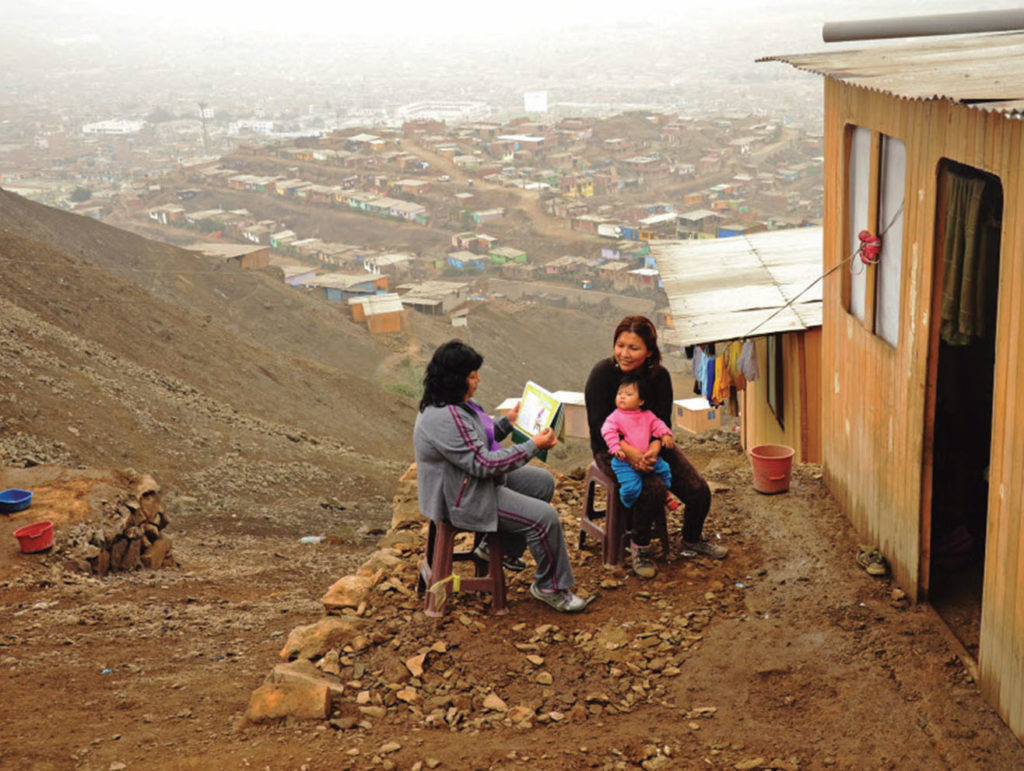 Community health worker sitting with a mother and young child outside a home on a Peruvian hillside,with homes visible in the distance