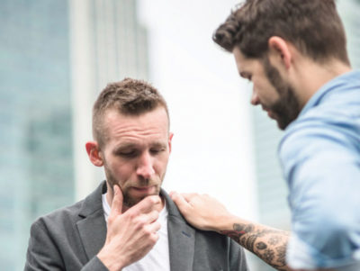 Two men in serious discussion, one in a gray suit jacket and the other in a blue shirt with tattoos visible on his forearm