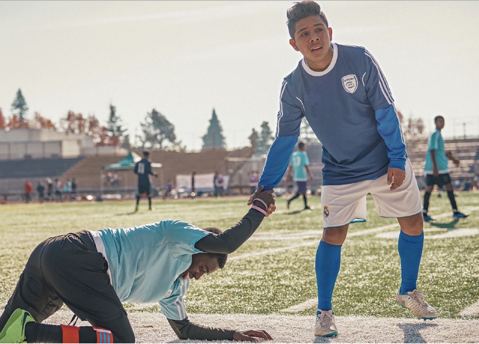 boy in soccer gear helping another boy stand up from where he's fallen on the soccer field