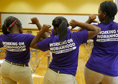 Three girls pointing to the back of their t-shirts which read "Working on Womanhood"