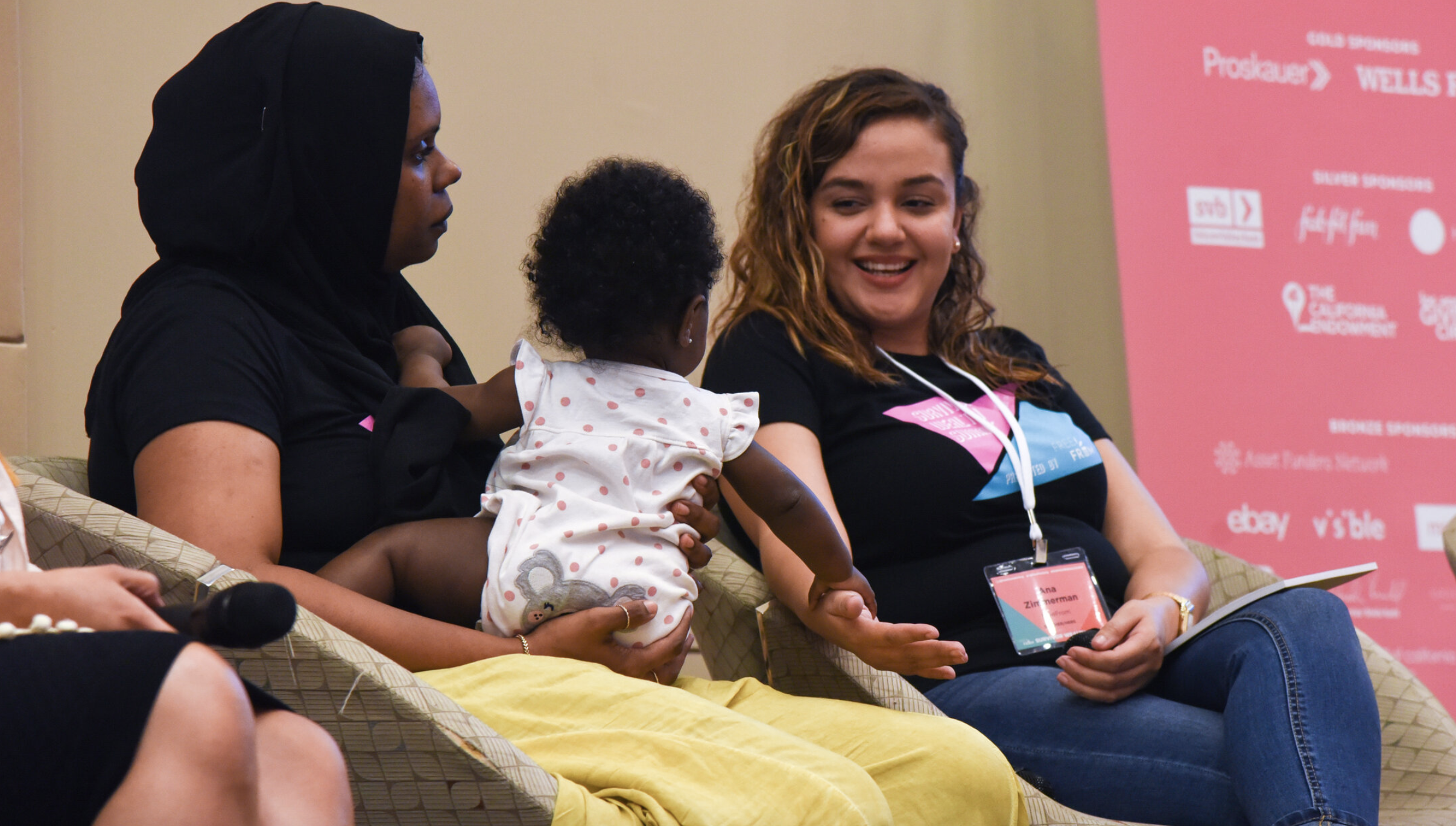 FreeFrom Woman in black hijab holding baby in ruffled onesie, sitting on dais with another curly-haired smiling woman in jeans, tee, and conference badge
