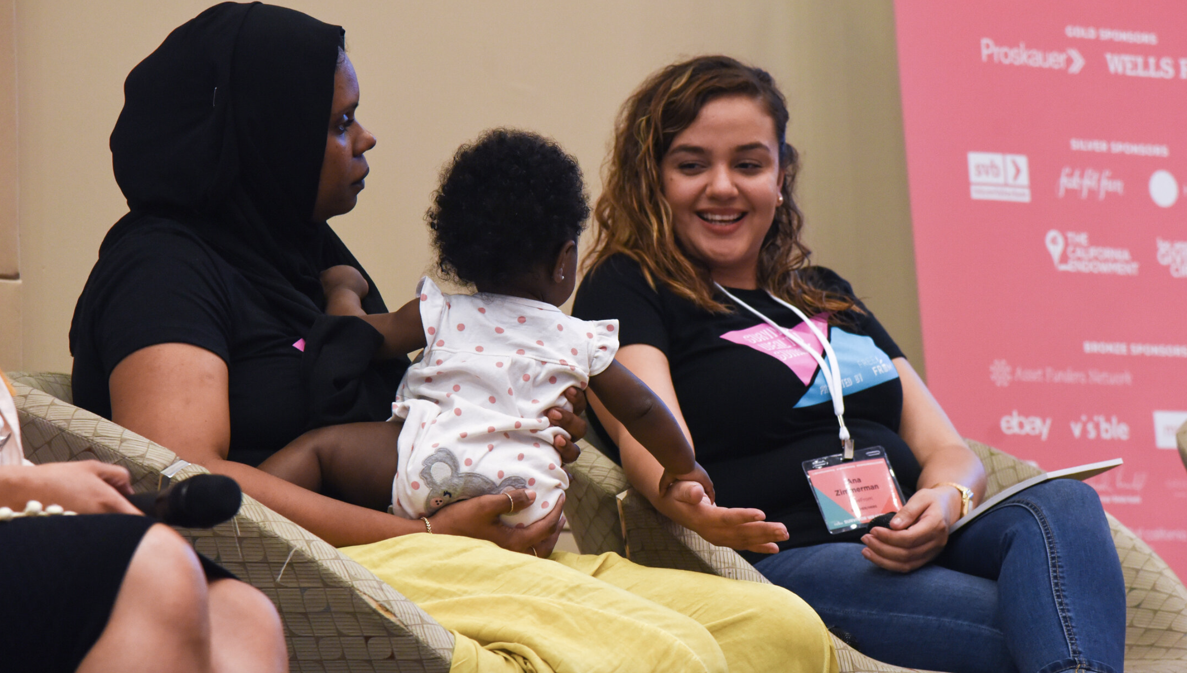 Woman in black hijab holding baby in ruffled onesie, sitting on dais with another curly-haired smiling woman in jeans, tee, and conference badge