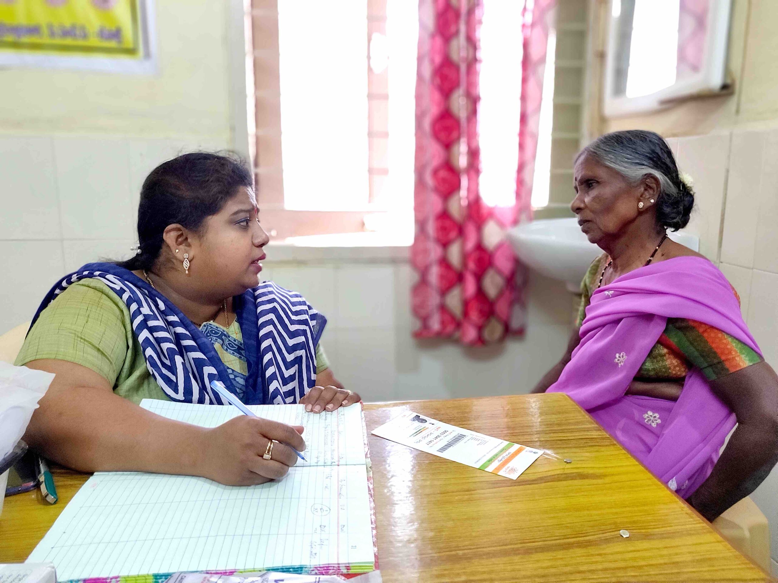 Two women sitting at a table looking at each other. One of the women has a pad of paper and a pen.