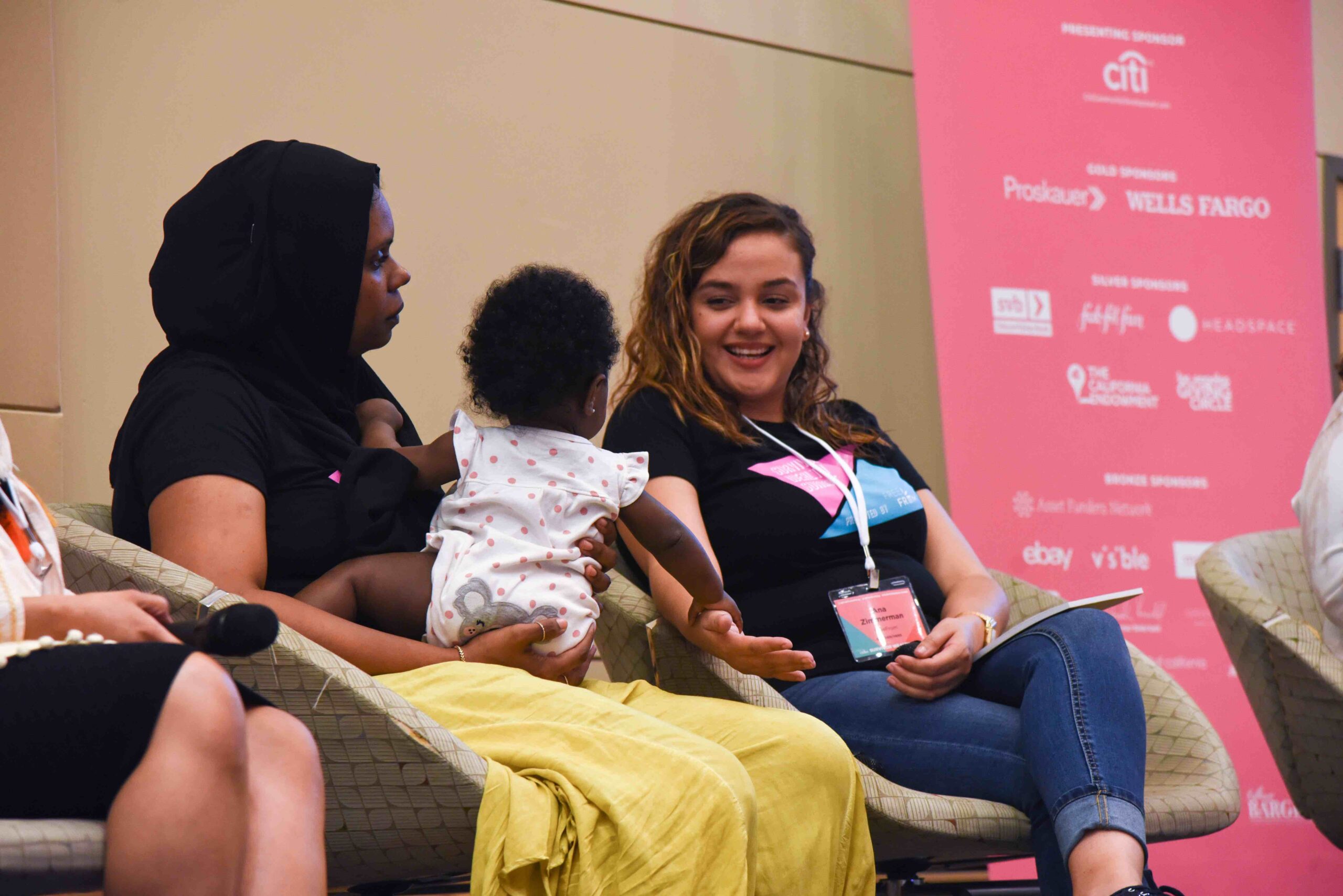 Two women sit next to each other at a conference. One woman has a baby on her lap.