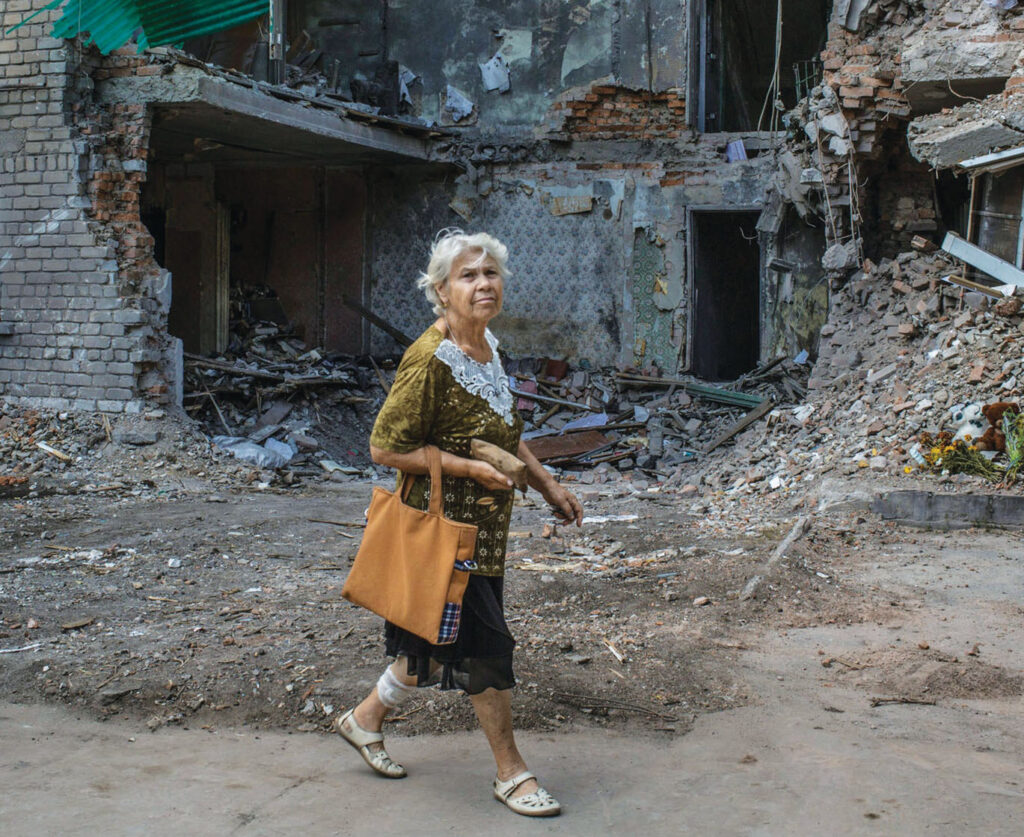 White-haired woman with a shopping tote walking past a crumbled and destroyed building