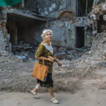 White-haired woman with a shopping tote walking past a crumbled and destroyed building
