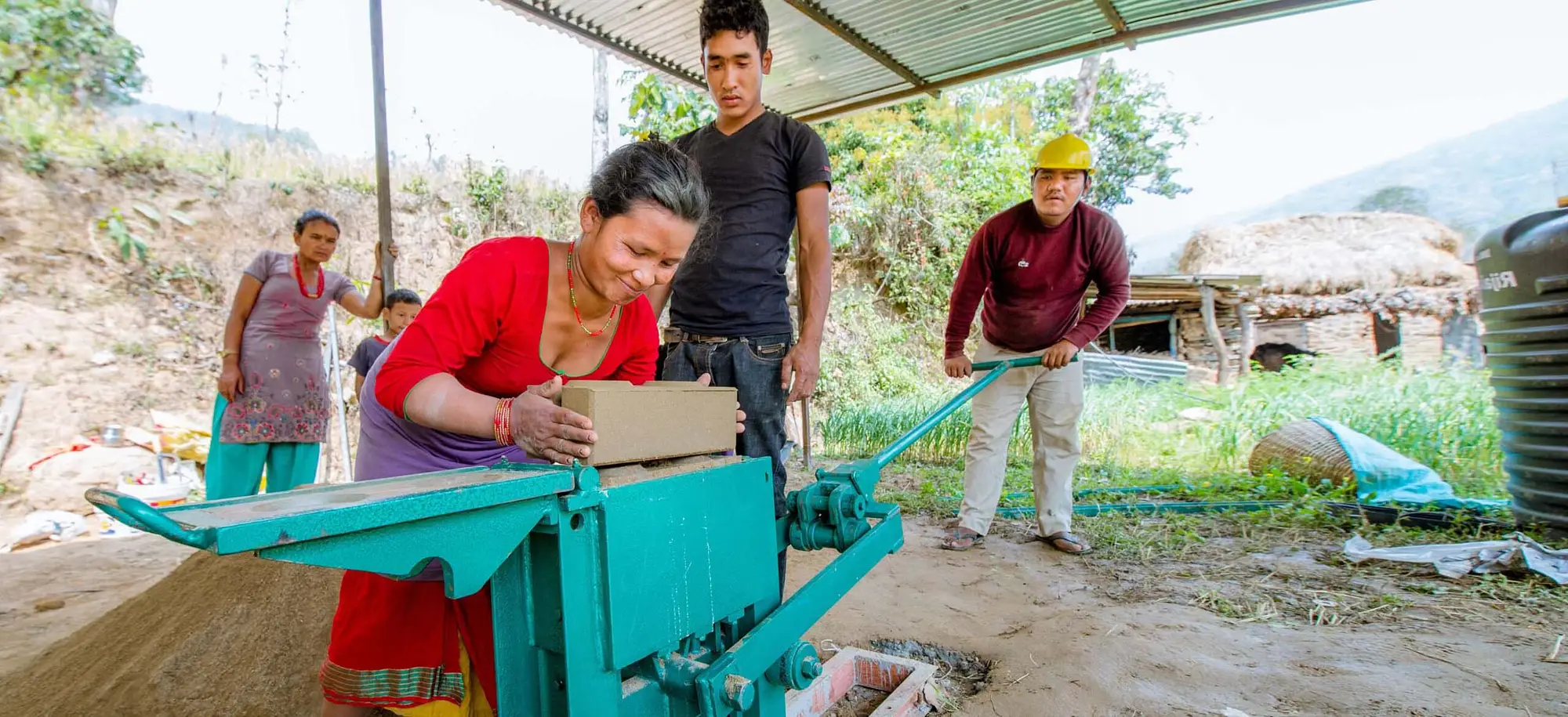 A woman makes a brick using Build Up Nepal's CSEB brick-making machine. A group of people look on, some in hard hats. 