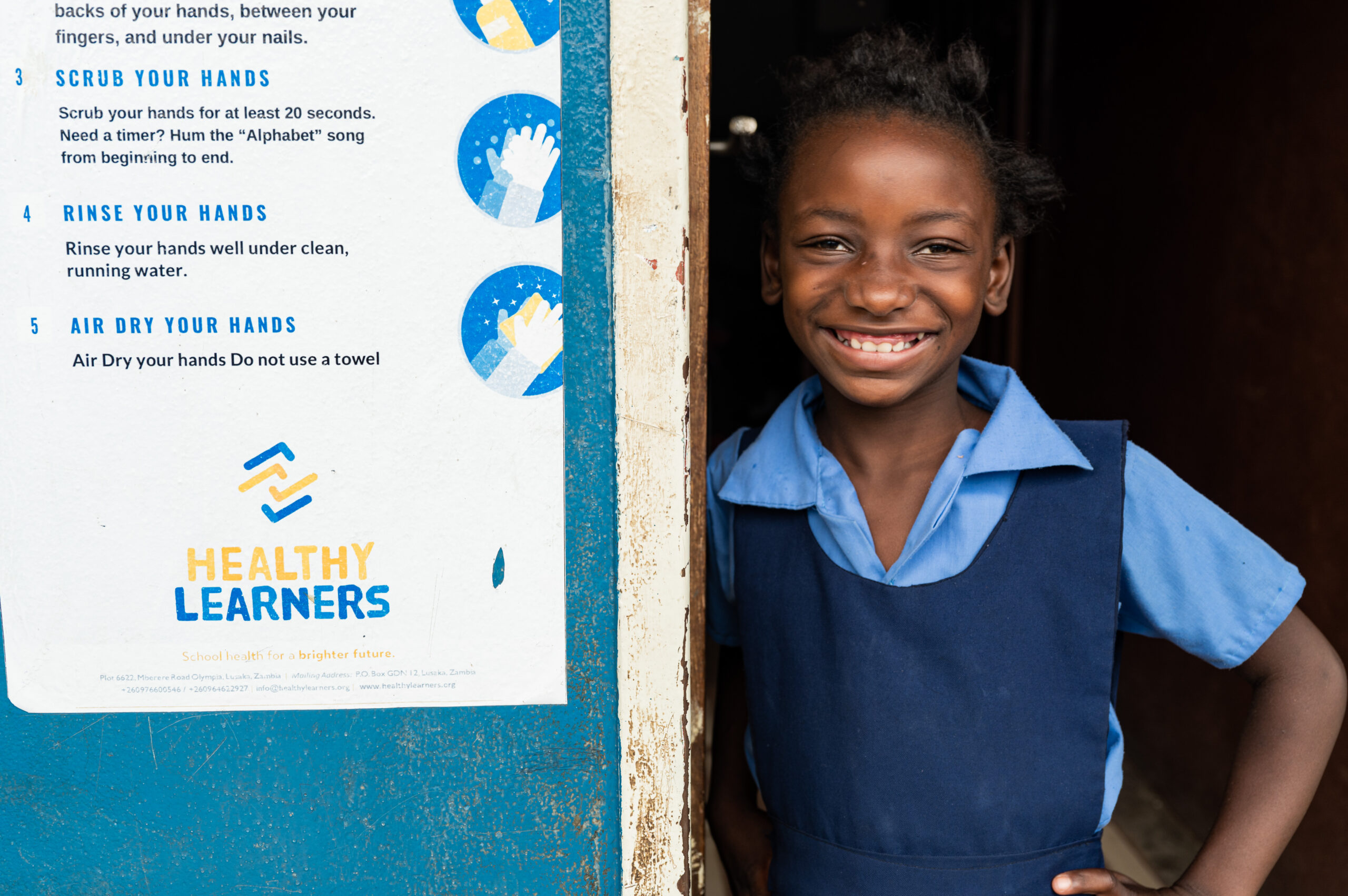 A young girl smiles next to a Healthy Learner's sign detailing how to properly wash your hands.