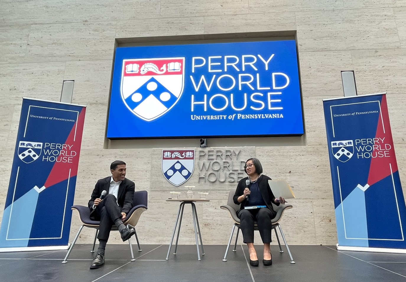 Rajiv Shah and Kat Rosqueta sit on a stage in front of a sign that says "Perry World House"